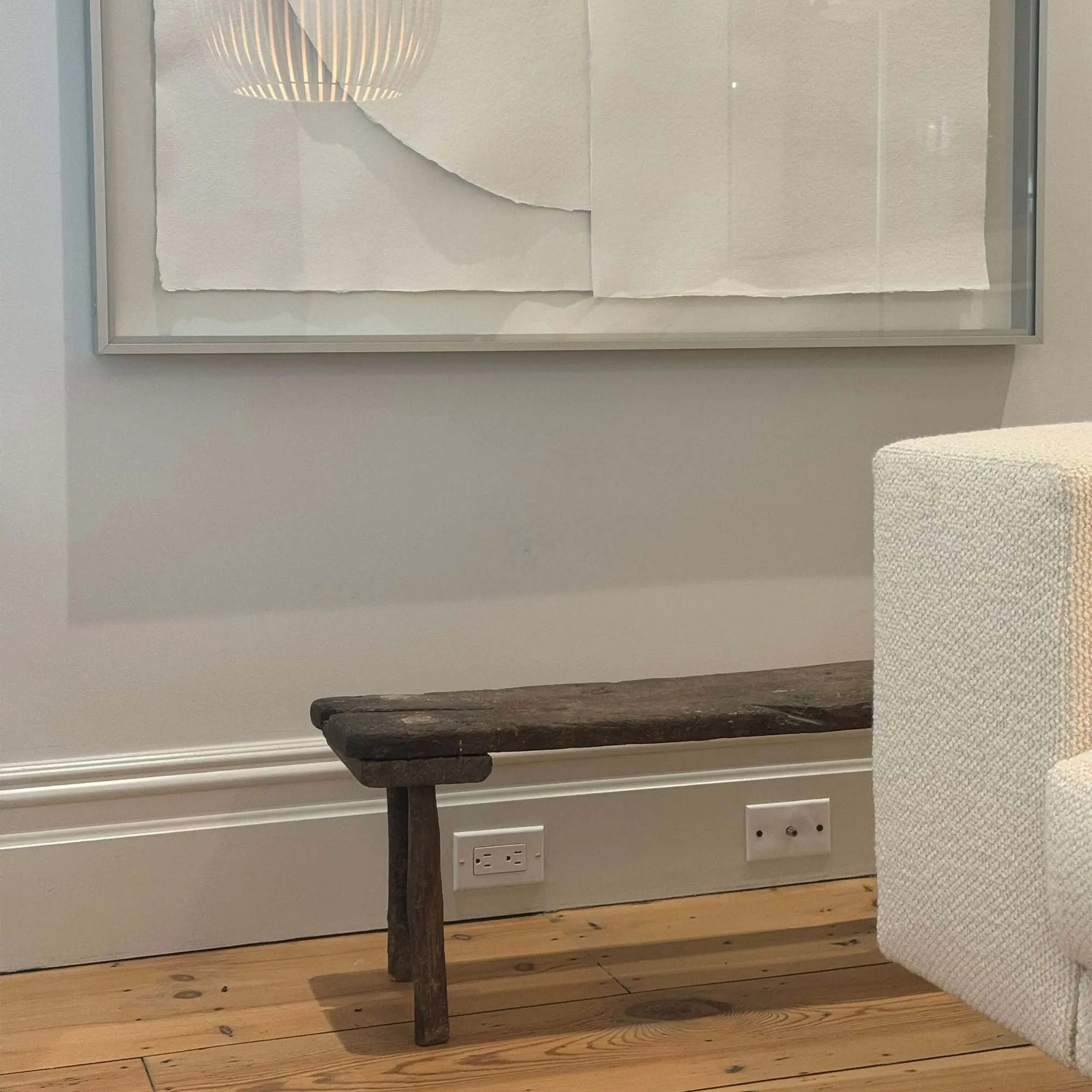 Partial side view of antique small table made of dark brown wood in a room with white background and hardwood floors. An all white framed work of art with the reflection of a round, white light fixture rests on the background wall behind the table. A white, woven textured couch sits partially in front of the table.